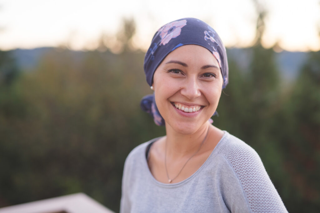 Beautiful Woman with Cancer Smiles A beautiful young woman wearing a head wrap looks toward the camera and smiles radiantly. She is standing outdoors and there are mountains and trees in the background.