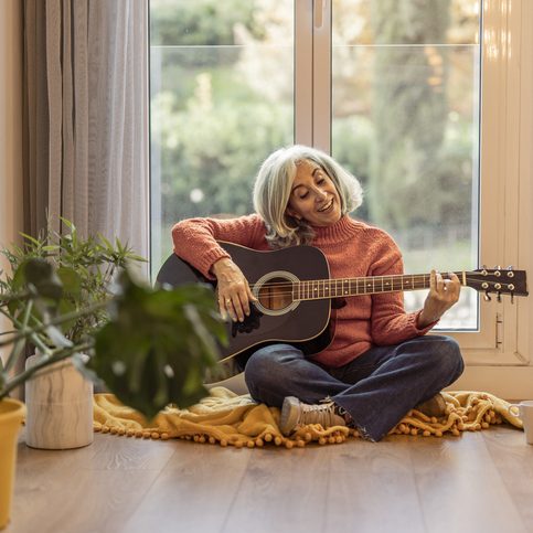 Senior woman playing guitar at home, grey-haired mature woman has fun at home by the window. life and home
